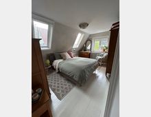 Schlafzimmer in einem Dachgeschoss mit schräge Wänden, einem Bett mit grauer Bettwäsche in der Mitte, Holzfußboden und einem gemusterten Teppich. Links und rechts befinden sich Holzmöbel, inklusive eines Spiegels und einer Kommode, sowie Fenster mit Blick nach draußen.