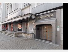 Straßenansicht eines grauen Gebäudes mit mehreren Eingängen. Links befinden sich Schaufenster mit Spielhallenwerbung und rechts ein Türbereich mit der Aufschrift „Insomnia“ über goldverzierten Doppeltüren.