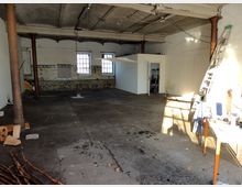 Alter, leerstehender Raum mit rauem Betonboden, unverputzten Wänden und teilweise freiliegender Backstein- und Fliesenstruktur. Der Raum hat sichtbare Eisensäulen, einfache Fenster mit Gitter und eine kleine, abgetrennte weiße Kabine auf der rechten Seite.