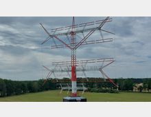 Eine große rote und weiße Sendeantenne mit komplexer Gitterstruktur befindet sich auf einer weitläufigen, grünen Wiese, umgeben von Bäumen. Der Himmel ist bewölkt, und im Hintergrund sind windkraftähnliche Strukturen erkennbar.
