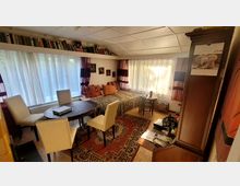 Gemütlich eingerichtetes Wohnzimmer mit einem runden Esstisch und vier weißen Stühlen, einer Tagesliege mit Kissen und einem kleinen Tisch daneben. Große Fenster mit weißen Vorhängen, Bücherregale an der Wand und ein Teppich auf dem Holzboden prägen den Raum.