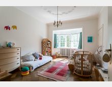 Helles Kinderzimmer mit Parkettboden, einer niedrigen Liegefläche, einem runden Holzgitterbett und einem Spielregal in Hausform. Ein großes Erkerfenster mit weißen Rahmen und grünen Vorhängen lässt Tageslicht herein, während Spielzeug und Bücher im Raum verteilt sind.