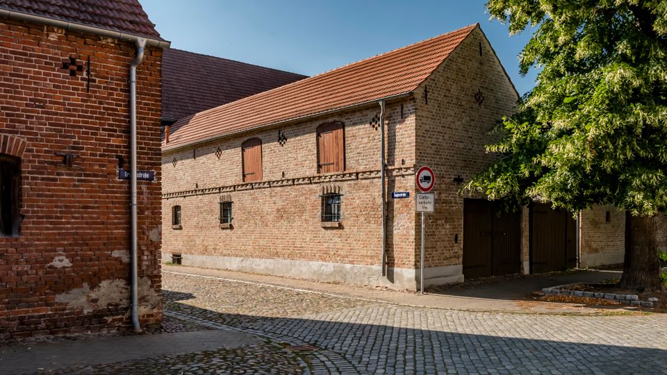 Kopfsteingepflasterte Straßenecke mit zwei historischen Backsteingebäuden mit Satteldächern, kleinen vergitterten Fenstern und hölzernen Klappläden. Rechts steht ein großer Baum am Gehweg neben Einfahrtstoren und einem Verkehrsschild.
