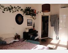 Helles Schlafzimmer mit Bett im Vordergrund, einem schwarzen Klavier an der Wand sowie Wandregalen mit Büchern und rankenden Zimmerpflanzen. Neben einer offenen Tür stehen ein großer weißer Kleiderschrank und ein Deckenlampenschirm; an der Wand hängen Dartscheibe und Bild, Sonnenlicht fällt auf den Holzboden.