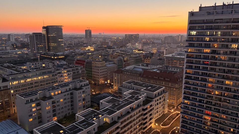 Stadtansicht bei Sonnenuntergang mit mehreren modernen und älteren Gebäuden, teilweise beleuchtet, sowie einem hohen Wohnhochhaus im Vordergrund rechts. Der Himmel zeigt einen orangefarbenen Streifen über der abklingenden Dämmerung.