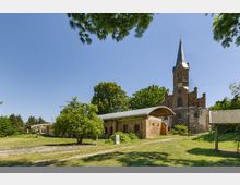 Kirchhof mit einer Backsteinkirche im gotischen Stil, deren Turm mit einer Uhr und einer Spitze mit Kreuz abschließt, umgeben von Bäumen und Wiesen; im Vordergrund ein Gebäude mit rotem Mauerwerk und modernem Metalldach.