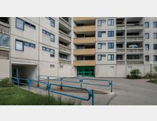 Wohnkomplex mit mehreren Etagen und Balkonen, teils in gelber und beige-weißer Fassade gestaltet, blaue Fensterrahmen und ein grüner Eingangsbereich. Im Vordergrund führt eine Rampe mit blauen Geländern über eine kleine Grünfläche.