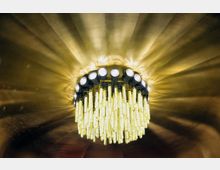 Ein Kronleuchter mit hängenden, leuchtenden Kristallketten, umgeben von einer goldfarbenen Decke, die strahlenförmige Lichtmuster reflektiert.