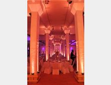 Großer Saal mit modernem, festlichem Ambiente, geprägt von hohen weißen Säulen, warmem, pink-orangen Licht und runden Tischen mit weißen Tischdecken und Stuhlhussen. Die Decke ist schlicht und mit kleinen Deckenstrahlern ausgestattet, während im Hintergrund eine Servierkraft sichtbar ist.