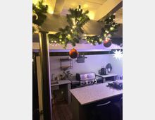 Außenküche mit überdachtem Bereich, geschmückt mit Tannengirlanden, Lichterketten und Kugeln. Ausstattung umfasst eine Stein-Arbeitsplatte, Edelstahlgrill, Spüle und dekorative Elemente wie Laternen und Pflanzen.