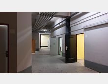 Langer, nüchterner Korridor in einem industriellen oder funktionalen Gebäude mit mehreren offenen Türen zu unterschiedlich beleuchteten Räumen. Sichtbare Rohrleitungen verlaufen unter der Decke, die Wände sind grau gestrichen, und der Boden besteht aus Beton.