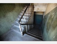 Schmales Treppenhaus in einem älteren Gebäude mit abgenutzten Holzstufen und einem schwarzen Handlauf mit weißen, abgeplatzten Geländerstäben. Die Wände sind zweifarbig grün und beige gestrichen und zeigen starke Gebrauchsspuren sowie abgeblätterte Farbe.