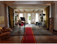 Industrieähnlicher Innenraum mit Einrichtung im Vintage-Stil, darunter Sessel, ein roter Teppichläufer und große Fenster mit hellem Tageslicht. Filmset-Ausstattung wie Beleuchtung und Kamerastative ist aufgebaut, und eine Person steht in der Mitte des Raums.