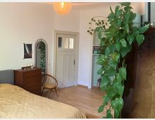 Schlafzimmer mit Holzfußboden, einem Bett mit gelber Tagesdecke, einer Kommode mit Kerzen, einem bogenförmigen Spiegel sowie einer Kletterpflanze, die von einem hohen Möbelstück hängt. Eine Tür führt zu einem angrenzenden Raum mit grünen Fliesen.