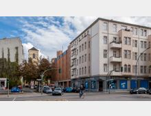 Straßenkreuzung in einem Stadtgebiet mit Wohn- und Geschäftsgebäuden. Links befindet sich ein Gebäude mit bewachsener Fassade, dahinter eine gelbe Kirchturmspitze, rechts ein Eckgebäude mit Balkonen und einer Reinigung im Erdgeschoss. Im Vordergrund Verkehr mit Autos und einem Fahrradfahrer.