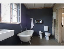 Ein Badezimmer mit dunkelgrauen Wänden, freistehender Badewanne, weißem Waschbecken, einer Toilette und einem kleinen weißen Regal an der Wand. Ein Fenster lässt Tageslicht herein, und die Backsteinwand ist neben der Tür sichtbar.