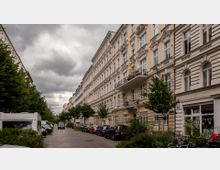 Straßenszene mit mehrstöckigen Altbauwohnhäusern in hellen Farben und aufwendiger Fassadengestaltung, links und rechts gesäumt von kleinen Bäumen. Am Straßenrand sind parkende Autos und ein Wohnwagen zu sehen, der Himmel ist bewölkt.