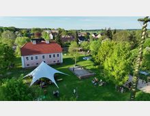 Luftaufnahme eines grünen Gartengeländes mit einem rosa Gebäude mit rotem Ziegeldach im Zentrum. Im Vordergrund ein weißes Zelt mit Sitzgruppen darunter, umgeben von Bäumen, Rasenflächen und kleinen Gehwegen. Im Hintergrund sind Wohnhäuser einer ländlichen Umgebung zu sehen.
