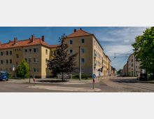 Straßenkreuzung in einem Wohngebiet mit mehrstöckigen, gelb verputzten Gebäuden und roten Ziegeldächern. Vor den Häusern sind Bäume, Laternen und geparkte Autos zu sehen, während Oberleitungen die Straße überspannen.