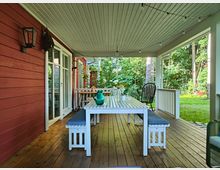 Überdachte Veranda eines roten Holzhauses mit hellem Holzboden, einem weißen Tisch mit Bänken und Gartenstühlen. Im Hintergrund befinden sich ein grüner Garten und Bäume, sowie Lichterketten und Wandleuchten als Dekoration.