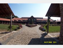 Gepflasterter Weg führt zu einem Gebäudeensemble mit roten Ziegeldächern und einer Fassade mit Giebeln; umgeben von Blumenbeeten und einer Grünanlage. Holz- und Backsteinkonstruktionen rahmen den Eingangsbereich.