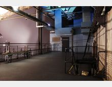 Industrieller Innenraum mit hohen Decken, sichtbaren Rohren, Metallgerüsten und einer Treppe, die in ein tiefer gelegenes Stockwerk führt. Die Wand links ist violett gestrichen, und rechts ist eine Betonwand mit Geländern und abgesperrten Bereichen.