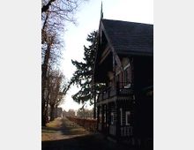Holzhaus mit verzierten Balken und einem steilen Giebeldach, flankiert von einer Allee mit kahlen Bäumen im Winter. Ein ruhiger Weg zieht sich entlang des Gebäudes, mit kahlen Hecken und vereinzelten Sitzbänken.