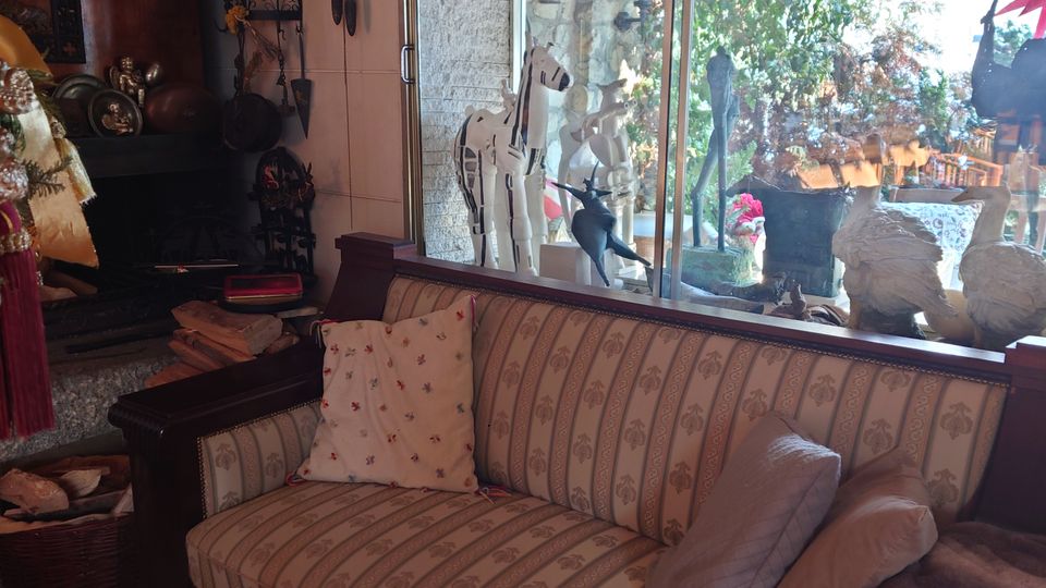 Gemütlicher Wohnraum mit einem gestreiften Sofa, dekoriert mit Kissen und einer Decke, vor einer großen Glasfensterfront. Im Hintergrund ist ein Außenbereich mit Pflanzen und Steinmauern sichtbar; nahe der Fensterbank stehen kleine Dekorationsgegenstände wie Holzspielzeug.