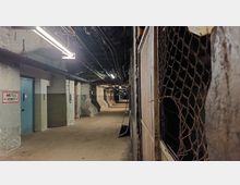 Langer, schmaler Kellerflur mit abgenutzten Betonwänden, freiliegenden Rohren und Kabeln an der Decke. Rechts befinden sich Drahtzäune, links Türen, und an der Wand hängt ein Schild mit der Aufschrift „Abstellen Verbot“.