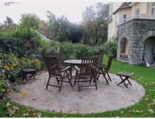 Garten mit einer gepflasterten Terrasse, auf der ein runder Holztisch und mehrere Holzstühle stehen, umgeben von Blumenbeeten, Büschen und einer Rasenfläche; im Hintergrund ein helles Gebäude mit einem Steinsockel und Bogendurchgang.