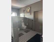 Badezimmer mit grau gefliesten Wänden und einem farbigen, geometrisch gemusterten Boden. Es gibt eine wandmontierte Toilette, ein Bidet, einen kleinen Spiegel am Fenster, sowie ein farbenfrohes Gemälde einer Frau an der Wand.