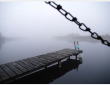 Holzsteg ragt in einen nebligen See, umgeben von Schilf am Ufer; im Vordergrund eine Kette und am Stegende ein kleiner Stuhl mit einer Laterne.