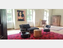 Heller Wohnraum mit großen Fenstern, eingerichtet mit schwarzen Sesseln, einem grauen Sofa, einer gelben Stehlampe und einem kleinen Holztisch, auf einem roten Orientteppich. An der Wand hängen ein großformatiges Porträt und ein antiker Holzkleiderschrank steht im Hintergrund.