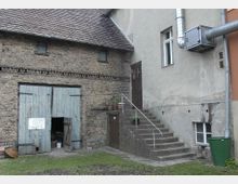 Backsteingebäude mit bläulich-grauer zweiflügeliger Holztür, über der ein Schild mit der Aufschrift „Ausfahrt freihalten“ hängt. Rechts davon befindet sich eine gemauerte Außentreppe mit Geländer, die zu einer braunen Tür in einem angrenzenden Putzbau führt.