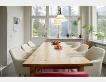 Heller Essbereich mit einem langen Holztisch, umgeben von weißen Polsterstühlen. Große Fenster im Hintergrund bieten Ausblick auf Bäume, und Pflanzen sowie Dekorationsobjekte stehen auf der Fensterbank.