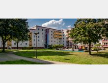 Mehrstöckiges Wohngebäude mit hellen Fassaden und farbigen Balkonen, umgeben von einer grünen Parkanlage mit Bäumen, Rasenflächen, Gehwegen und einem kleinen Spielplatz.