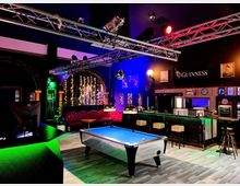 Innenraum einer Bar im Pub-Stil mit Billardtisch im Vordergrund, langer Theke mit Hockern und rotem Leder-Sitzbereich an der Rückwand. Über dem Raum hängen Metall-Traversen mit Bühnenleuchten; farbige Lichtakzente und Dekorationen wie Spiegelkugel und Guinness-Banner prägen die Atmosphäre.