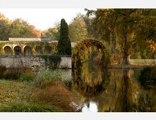 Ein historisches Gebäude mit einer Arkade aus Naturstein steht am Rand eines ruhigen Teichs, umgeben von herbstlich gefärbten Bäumen und Pflanzen. Ein bogenförmiges Rankgitter mit buntem Laub spannt sich über den Teich.