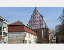 Straßenansicht eines Kirchengebäudes mit großer, dreieckiger Giebelfassade, die mit einem rot umrahmten Bogenmuster verziert ist, sowie einem seitlichen Backsteinbau mit hohen Rundbogenfenstern. Im Vordergrund stehen Wohnhäuser mit roten Ziegeldächern, Straßenlaternen und Verkehrsschildern, daneben Bäume entlang der Straße.