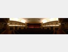 Ein klassischer Kinosaal mit Reihen dunkler Kinositze, einer Bühne mit geschlossenem roten Vorhang und holzgetäfelten Wänden. Die Decke ist leicht gewölbt und diffus beleuchtet, wobei kleine Leuchten an den Wänden sichtbar sind.