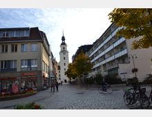 Gepflasterte Fußgängerzone mit einem Kirchturm im Hintergrund, flankiert von modernen Wohn- und Geschäftshäusern. Im Vordergrund sind Fahrradständer, Bäume mit Herbstlaub und Menschen zu Fuß oder mit dem Fahrrad zu sehen.