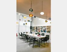 Großer, moderner Besprechungsraum mit einem langen Tisch, umgeben von schwarzen Stühlen. Im Hintergrund ein Regal mit dekorativen Glasobjekten und weiße Vorhänge; von der Decke hängen bunte, kugelförmige Leuchten in unterschiedlichen Höhen.
