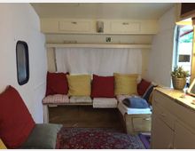 Wohnbereich in einem Wohnwagen mit Sitzbank, die mit roten, gelben und gestreiften Kissen dekoriert ist. Ein kleiner Teppich liegt auf dem Boden, und die Einrichtung umfasst weiße Wände, Hängeschränke und eine Arbeitsfläche mit einer Topfpflanze.