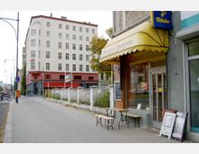 Straßenszene in einer Stadt mit einem Café mit gelbem Markisendach und Holzbänken auf dem Gehweg im Vordergrund. Im Hintergrund ist ein mehrstöckiges Wohn- und Geschäftsgebäude mit hellen und roten Fassaden sowie ein Gehweg mit Laternen und Fahrrädern zu sehen.