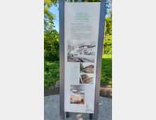 Informationstafel im Freien mit der Überschrift „Park und Schlösschen Monplaisir“. Die Tafel zeigt Text zur Geschichte des Ortes sowie historische und aktuelle Fotografien des Gebäudes. Sie steht auf einem gepflasterten Weg, umgeben von grünen Bäumen und Vegetation.