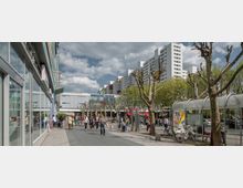 Städtischer Platz mit einem Einkaufszentrum im Hintergrund, beschriftet als „Märkisches Zentrum“. Links eine Glasfassade, rechts kahle Bäume und eine überdachte Bushaltestelle, dahinter mehrstöckige Wohngebäude und Fußgänger.