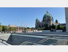 Blick auf den Berliner Dom mit seiner großen Kuppel und flankierenden Türmen, neben einer modernen Treppenabfahrt. Im Hintergrund sind weitere historische Gebäude sowie Bäume und Fahnen zu sehen.