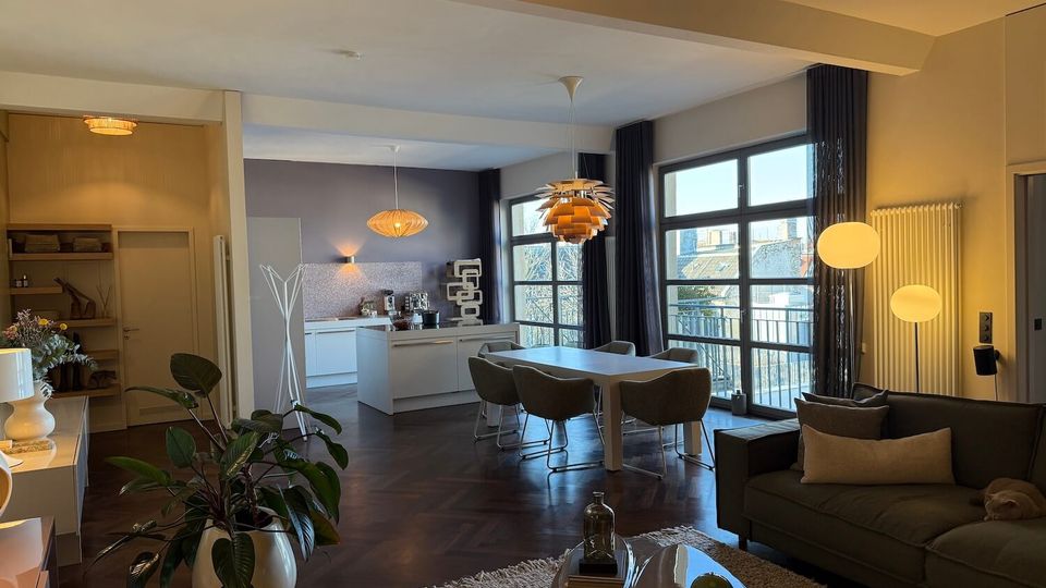 Open-plan modern living and dining room with a white kitchen island, pendant lights, and dark wood herringbone flooring. Large grid-style windows and glass doors with dark curtains open to a balcony, with a sofa and low coffee table in the foreground.