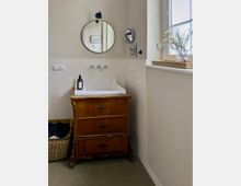 Helles Badezimmer mit wandhängendem Waschbecken auf einem antiken Holzschrank, runden Wandspiegeln und weißen Fliesen bis zur halben Wandhöhe. Rechts ein großes Fenster mit Fensterbank und Tablett mit Vase, darunter grauer Fliesenboden.