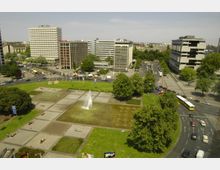 Stadtansicht mit einer großen Parkanlage im Vordergrund, die Wasserfontänen, Grünflächen und Gehwege umfasst. Im Hintergrund sind moderne Bürogebäude, eine Hauptstraße mit Autos sowie weitere Grünflächen sichtbar.